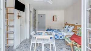 Ein kleines Schlafzimmer mit Bett, Tisch und Stuhl. Ein Deckenventilator und ein an der Wand befestigtes Fernsehgerät sind vorhanden.