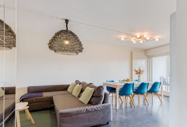 Una sala moderna con un sofá modular, una mesa de comedor con sillas azules y dos lámparas colgantes.