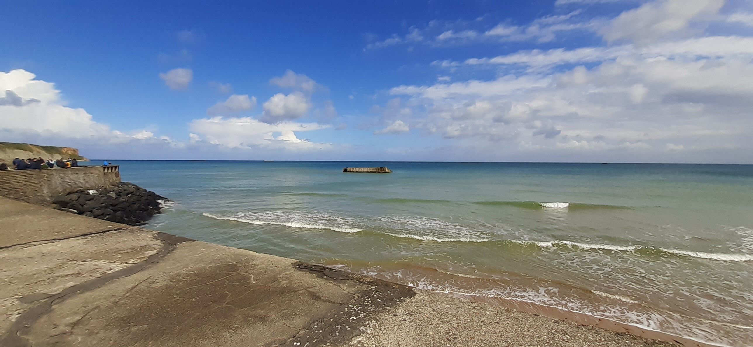A coastal scene with a concrete walkway, calm sea, and a distant pier.