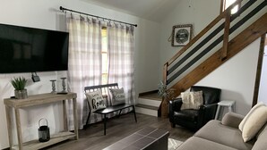 Una sala de estar con sofá, mesa de centro, televisor montado en la pared, escalera y ventana con cortinas.
