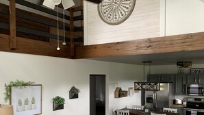 Amplia sala de estar con techo alto, ventilador de techo, mesa de comedor con sillas, cocina con electrodomésticos modernos y un rincón de descanso acogedor con sofá y reposapiés.