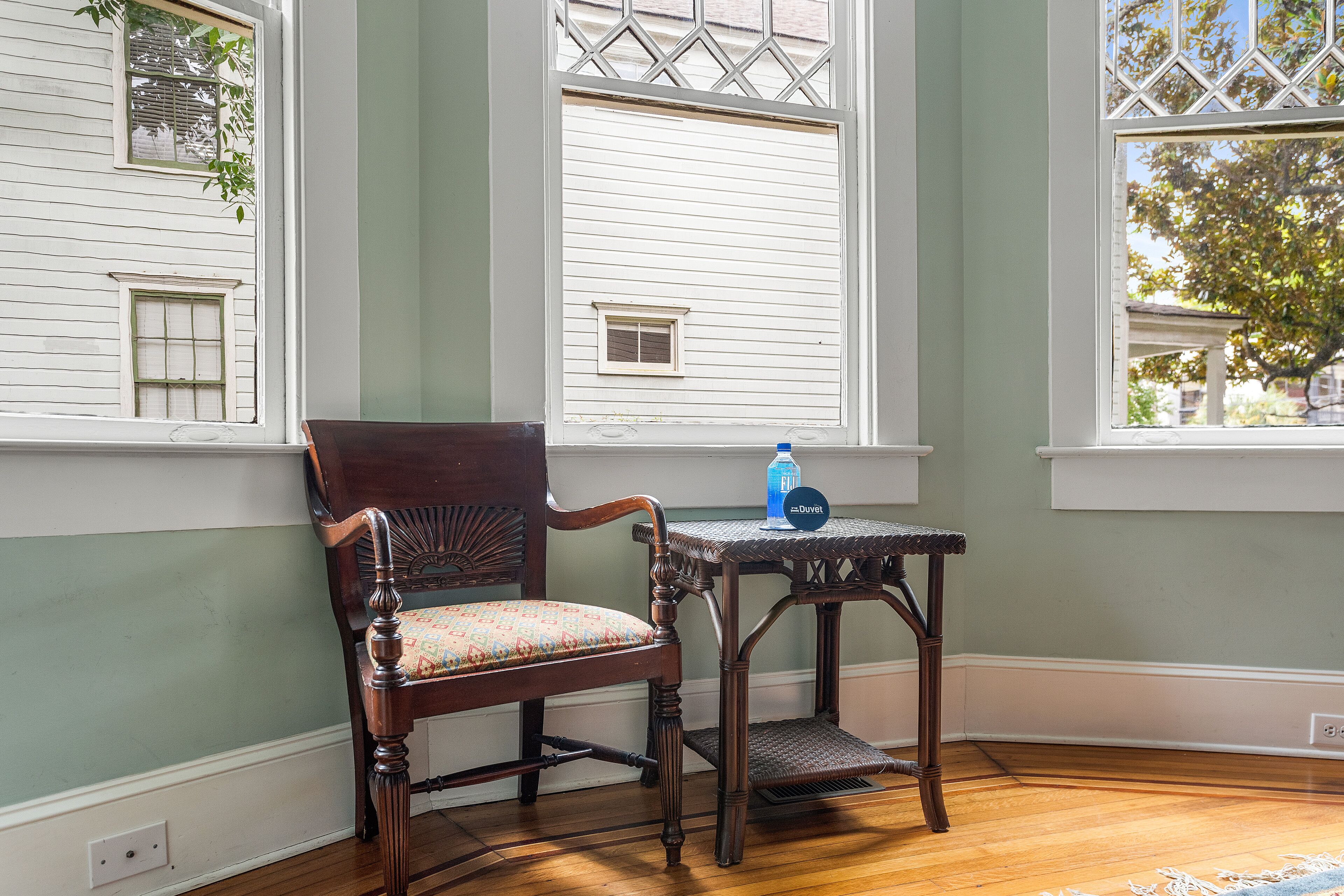 A room with a wooden chair, a small table with a bottle, and a window with a view of trees.
