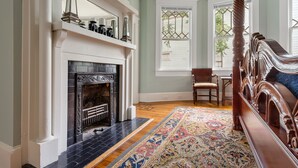 A room with a fireplace, a large window with a view, a wooden bed, and a patterned rug. - Downtown Luxury | Rutledge Avenue Inn (Suite A) (Charleston)