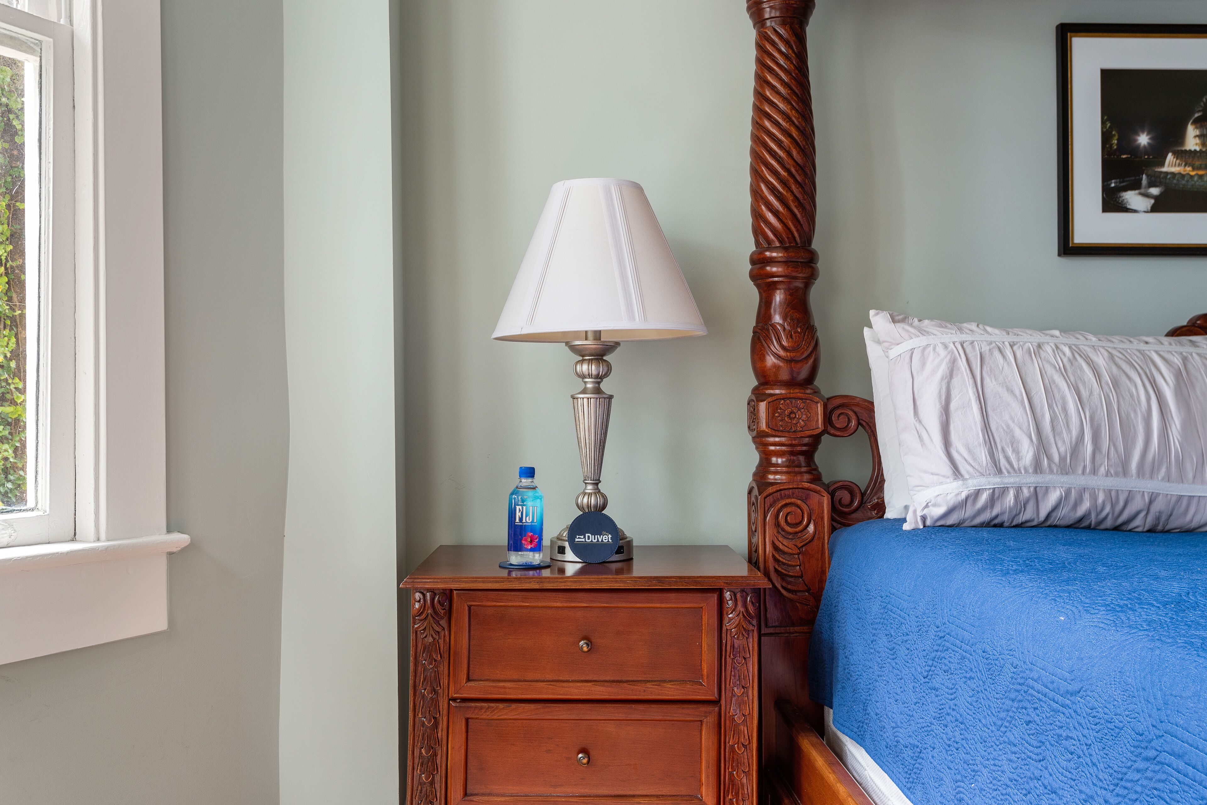 A bedroom with a wooden four-poster bed, a nightstand with a lamp, a bottle of Fiji water, and a framed picture on the wall.