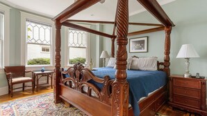 A four-poster canopy bed with a blue bedspread, a wooden nightstand with a lamp, and a patterned rug. - Downtown Luxury | Rutledge Avenue Inn (Suite A) (Charleston)