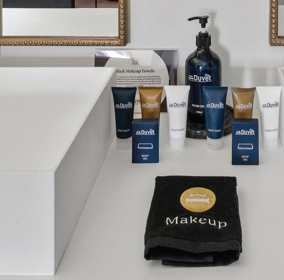 En badeværelsesvask med et spejl, et sort makeuphåndklæde og forskellige DuVet plejeprodukter, herunder shower gel, sæbe og balsam.