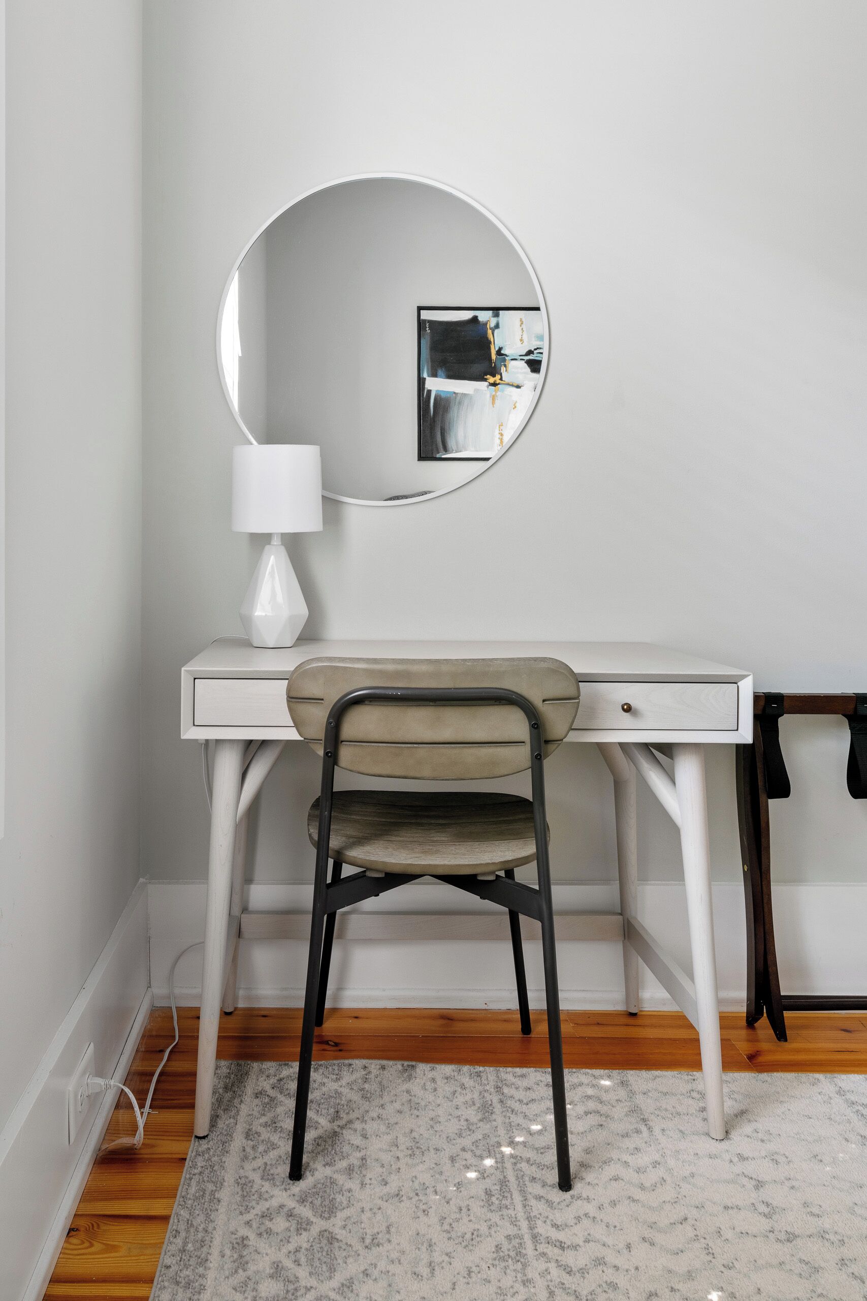 A white desk with a round mirror, a lamp, and a chair in a room with wooden flooring and a patterned rug.
