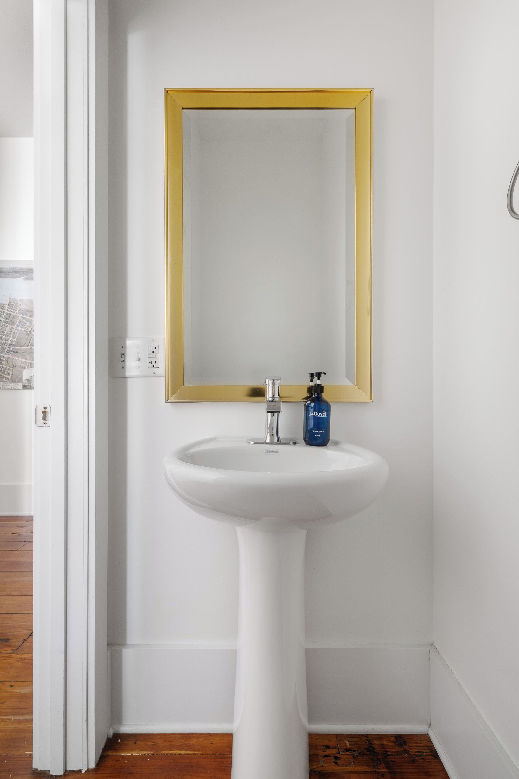 A white bathroom with a pedestal sink, a gold-framed mirror, and a blue bottle of hand soap.