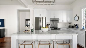 A modern kitchen with white cabinetry, a stainless steel refrigerator, a built-in oven, and a large island with bar stools. - Luxurious Escape in Downtown Charleston | Apollo (Charleston)
