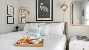 A neatly made bed with pillows, a tray with breakfast items, a framed bird artwork, wall-mounted lamps, and a mirror with a decorative frame. - Luxurious Escape in Downtown Charleston | Apollo (Charleston)