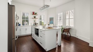 A modern kitchen with white cabinetry, a stainless steel refrigerator, and a central island with a built-in oven. - Luxurious Escape in Downtown Charleston | Apollo (Charleston)