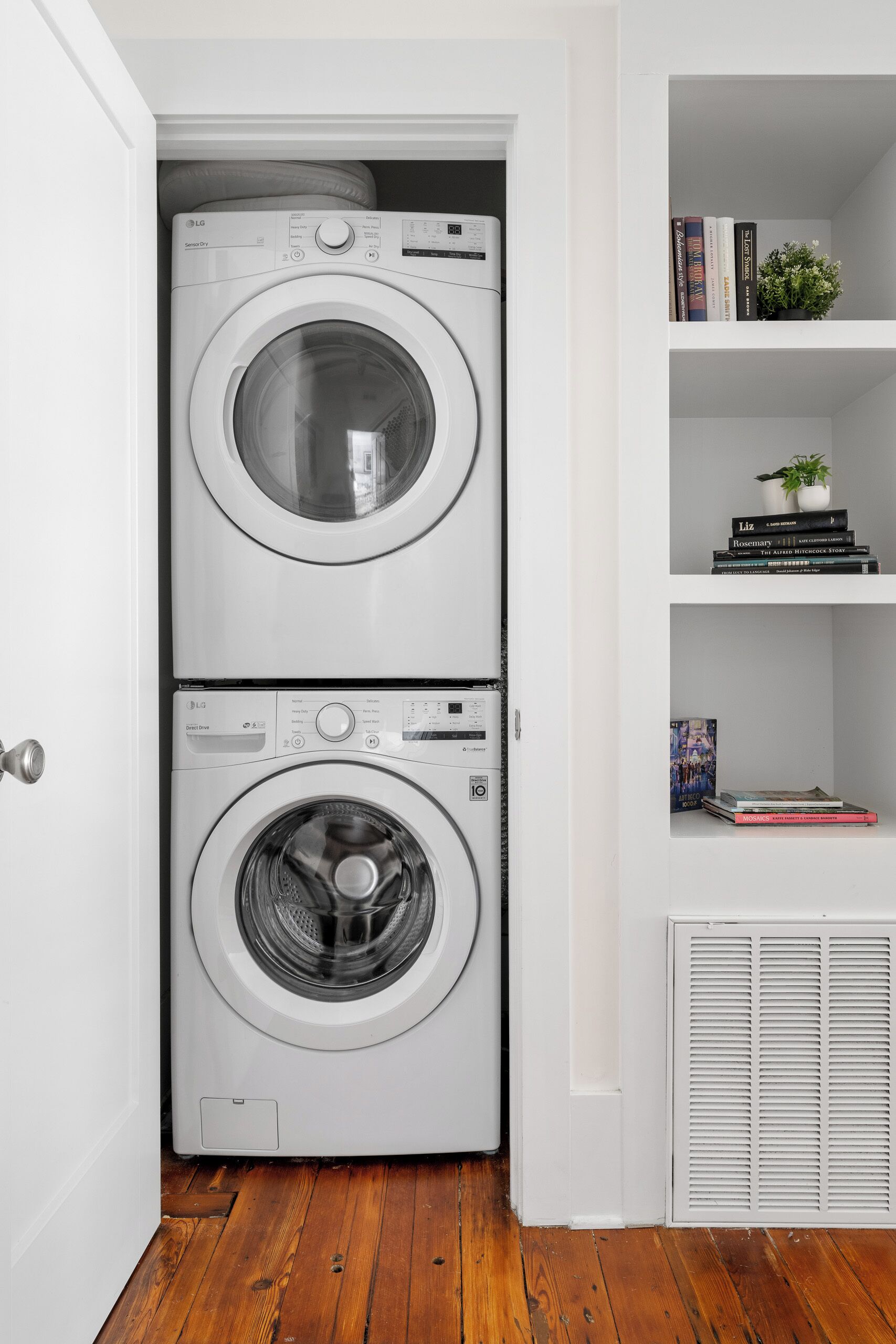 A laundry room with two stacked front-loading washing machines and a shelf with books and a small plant.