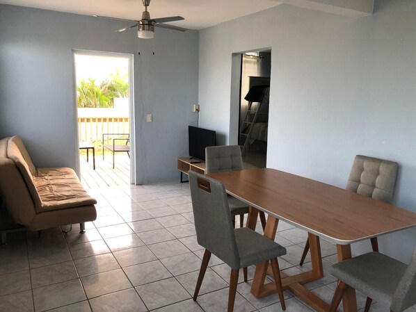 A dining area with a wooden table and four chairs, a sofa, a television, and a ceiling fan.