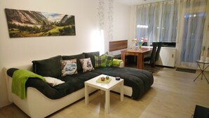Un salon avec un canapé noir et blanc, une petite table blanche sur laquelle se trouve un panier de fruits, un tableau représentant un paysage de montagne, une table à manger avec des chaises et une fenêtre avec des rideaux.