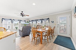 A dining area with a wooden table and chairs, a ceiling fan, and large windows with curtains. - Channel Cat - Gorgeous Cherry Grove Channel House with Pool (North Myrtle Beach)