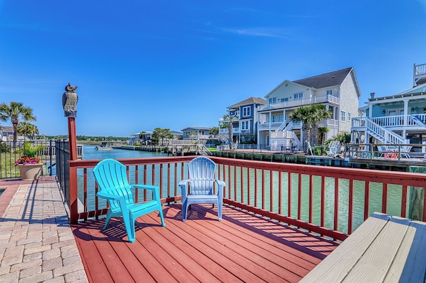 A wooden deck with two chairs, a railing, and a view of a water body and buildings. - Channel Cat - Gorgeous Cherry Grove Channel House with Pool (North Myrtle Beach)