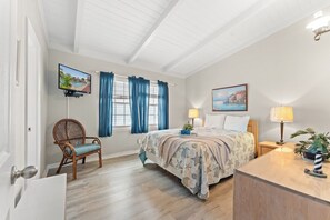 A bedroom with a bed, a chair, a desk, a TV, and a painting.