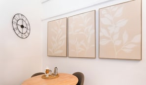 Uma área de refeições com uma mesa redonda, um relógio e três peças de arte com motivos de folhas.