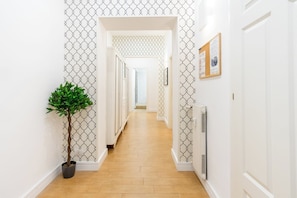Um corredor com papel de parede estampado, um vaso de planta, um radiador e uma porta com um mural de avisos.