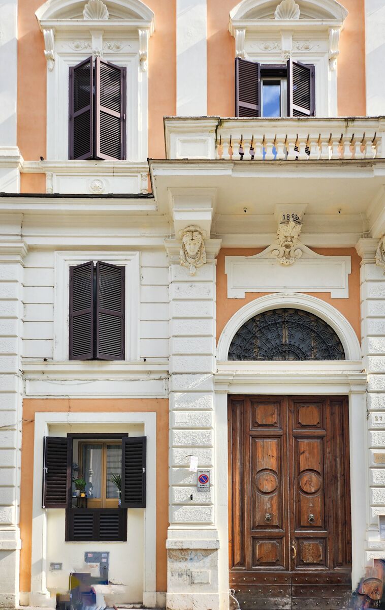 A building facade with a large wooden double door, a balcony with a railing, and a window with shutters.