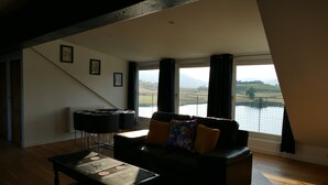 A living room with a sofa, a coffee table, and a piano. There are large windows with curtains, a view of a landscape, and a staircase leading to an upper level. - The Annexe at Loch View Farm will take your breath away (Crieff)