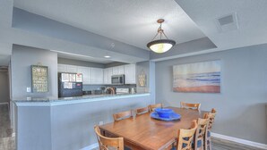 A dining area with a wooden table and chairs, a kitchen in the background, and a beach-themed painting on the wall. - Grand Atlantic 1901 - 4 Bedroom Ocean Front Condo (Myrtle Beach)