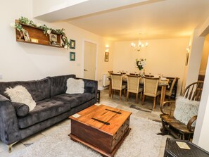 A living room with a dark grey sofa, a wooden coffee table, and a dining area with a wooden table and wicker chairs. - Stewart House (Whitby)
