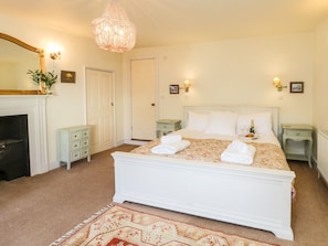 A bedroom with a large bed, two bedside tables, a chandelier, a fireplace, and a patterned rug. - Stewart House (Whitby)