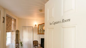 Une pièce avec une porte blanche portant l’inscription « Chambre du capitaine de navire », un miroir, une chaise et une petite table sur laquelle se trouve une plante.