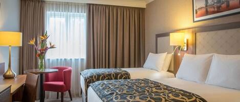 A hotel room with two beds, a desk, a chair, a vase with flowers, and a cityscape painting on the wall.