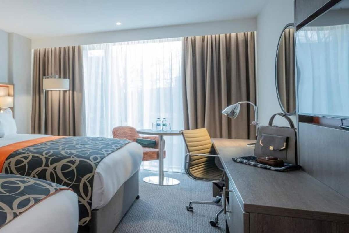 Habitación de hotel con dos camas, un escritorio, una silla y un ventanal con cortinas.