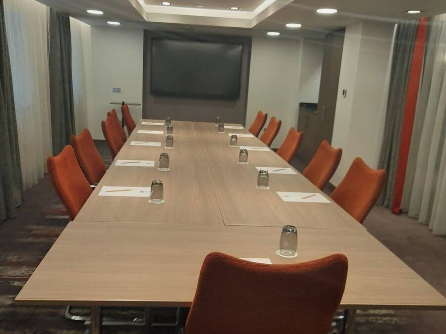 A conference room with a long table, orange chairs, and a large screen at the front.