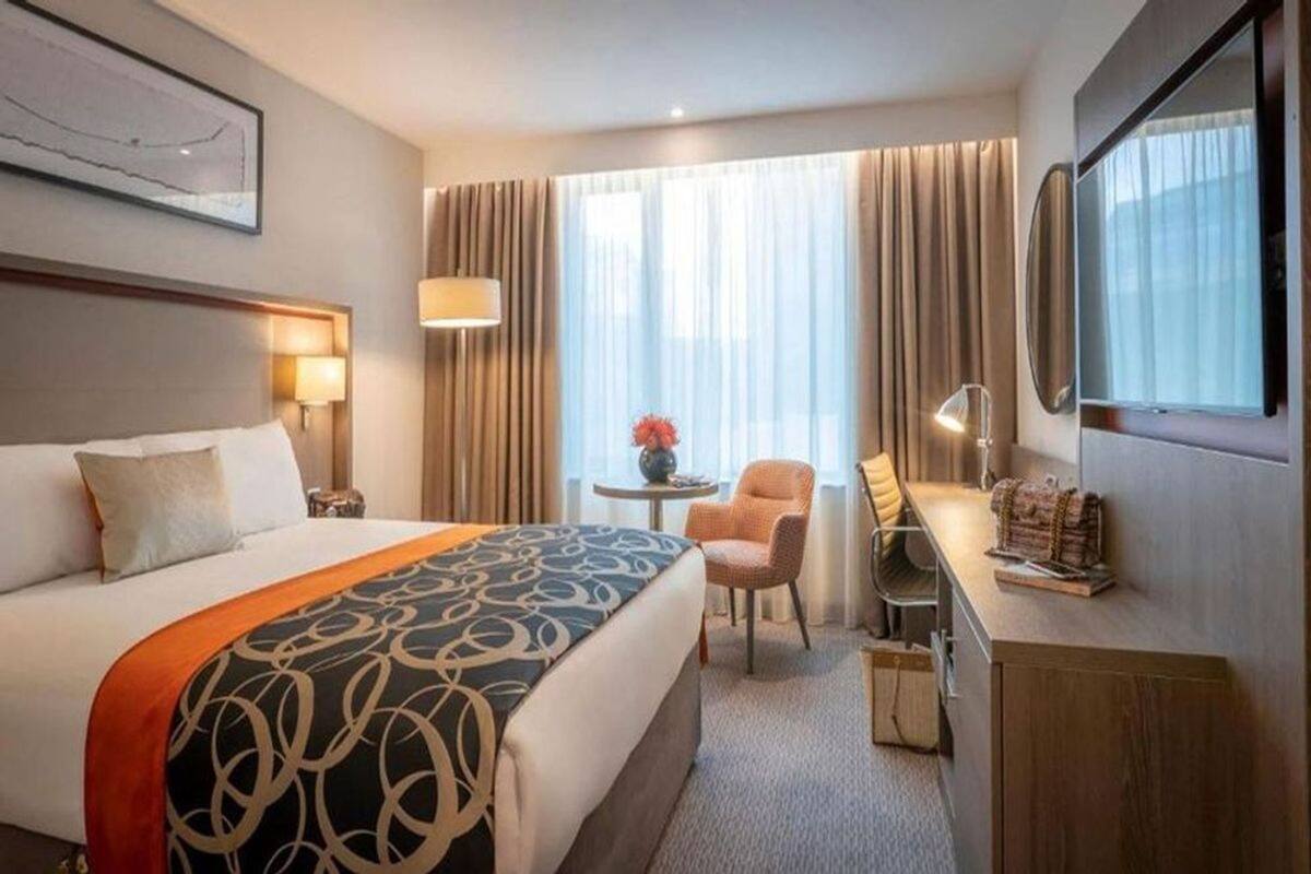 Habitación de hotel con una cama grande, un escritorio con silla, una mesita con un florero y un televisor.