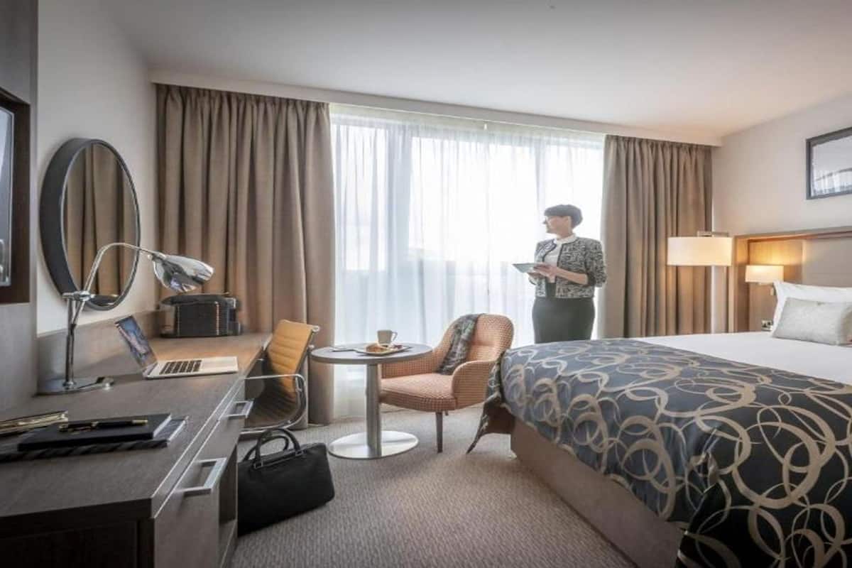 Habitación de hotel con una cama grande, un escritorio con lámpara, una silla, una mesita y una ventana con cortinas.