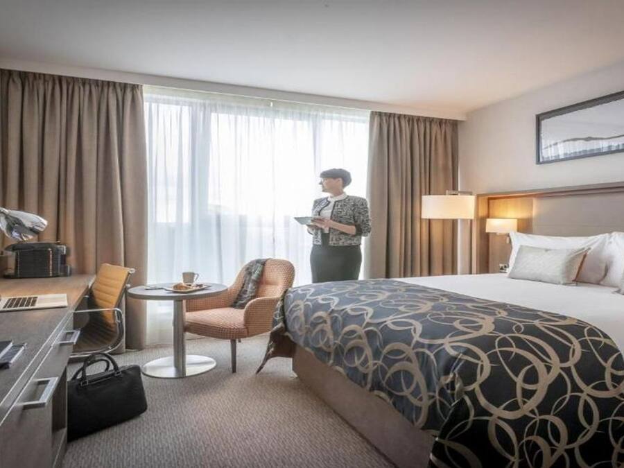 Quarto de hotel com cama, escrivaninha, cadeira, luminária e janela com cortinas.