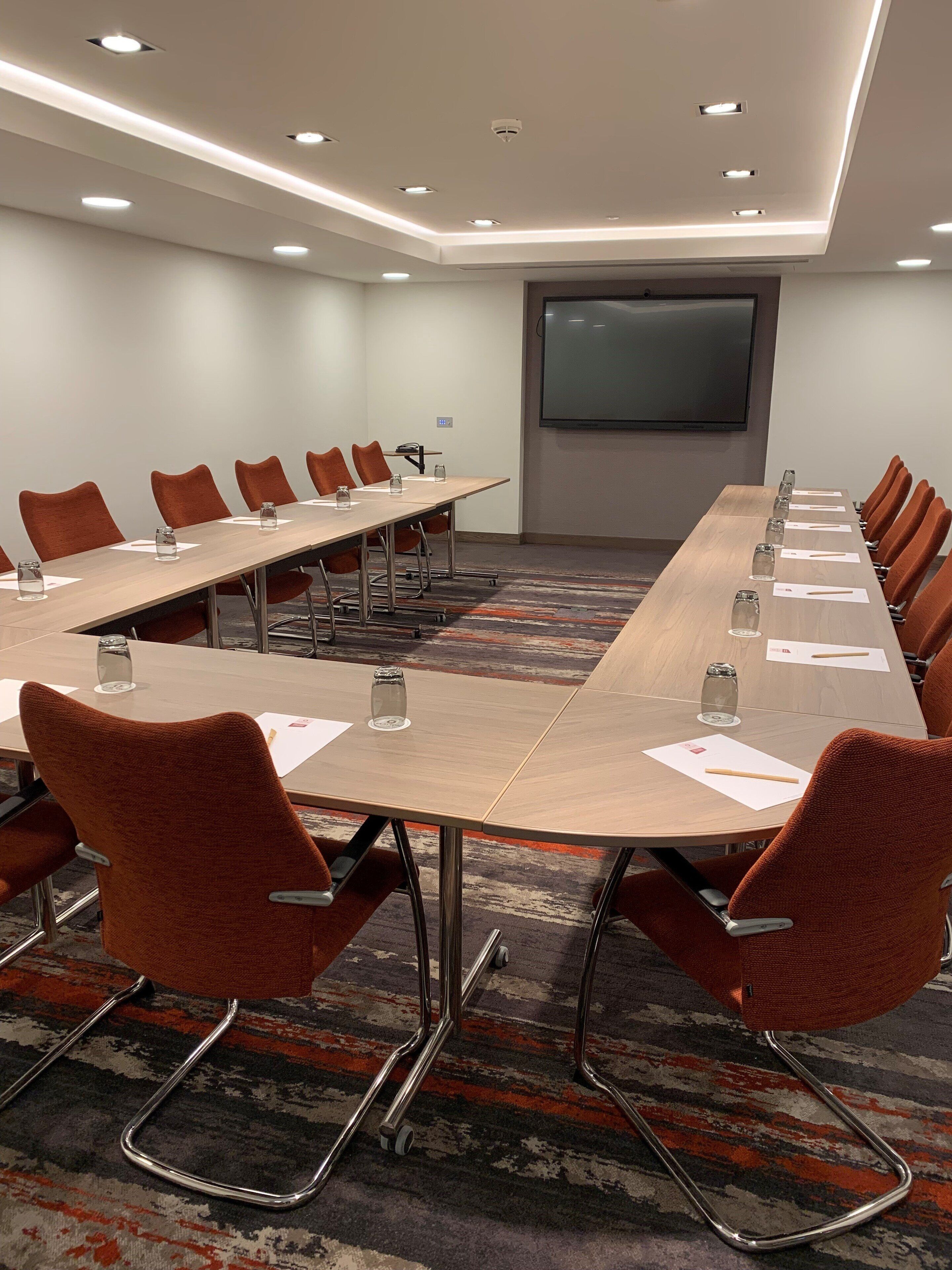 Une salle de conférence avec une longue table, des chaises orange et un écran de projection mural.
