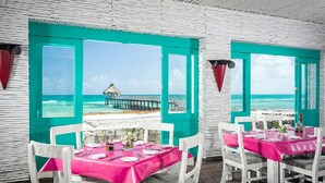 A dining area with turquoise walls and windows overlooking a beach and pier. Tables are set with pink tablecloths and white chairs. - Vidanta Mayan Palace 2 bedroom 2 Bath  (Riveria Maya)