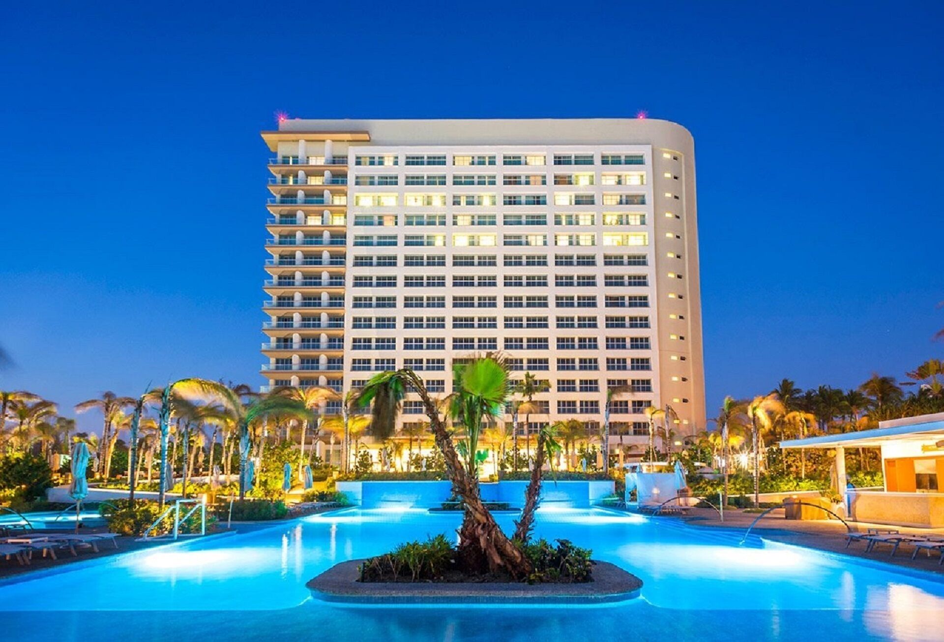 A hotel with a large pool, palm trees, and a well-lit building at dusk.
