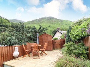 A patio with wooden furniture and a small shed, surrounded by lush greenery and hills in the background. - Capel Cottage (Penmaenmawr)