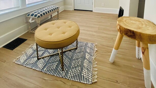A spacious room with large windows, a bench, a round ottoman, and a small wooden stool.