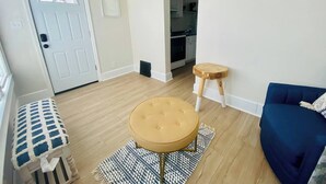 A living room with a blue sofa, a round stool, a patterned bench, and a door with a window.
