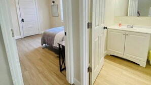 A bedroom with a bed, a bedside table, and a nightstand. There is a bathroom with a sink, mirror, and cabinet. The room has wooden flooring and white walls.