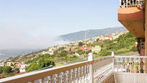 A balcony with a white balustrade overlooking a hillside town with houses and greenery.