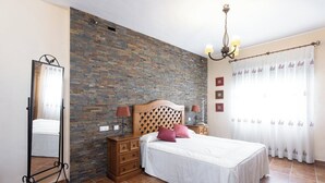 A bedroom with a brick wall, a bed with white bedding, two bedside tables with lamps, a mirror, and a window with curtains.