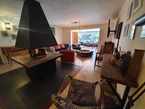 A spacious living room with a large black table, a red sofa, and a wicker chair. There is a wooden cabinet and a dining table with chairs in the background.