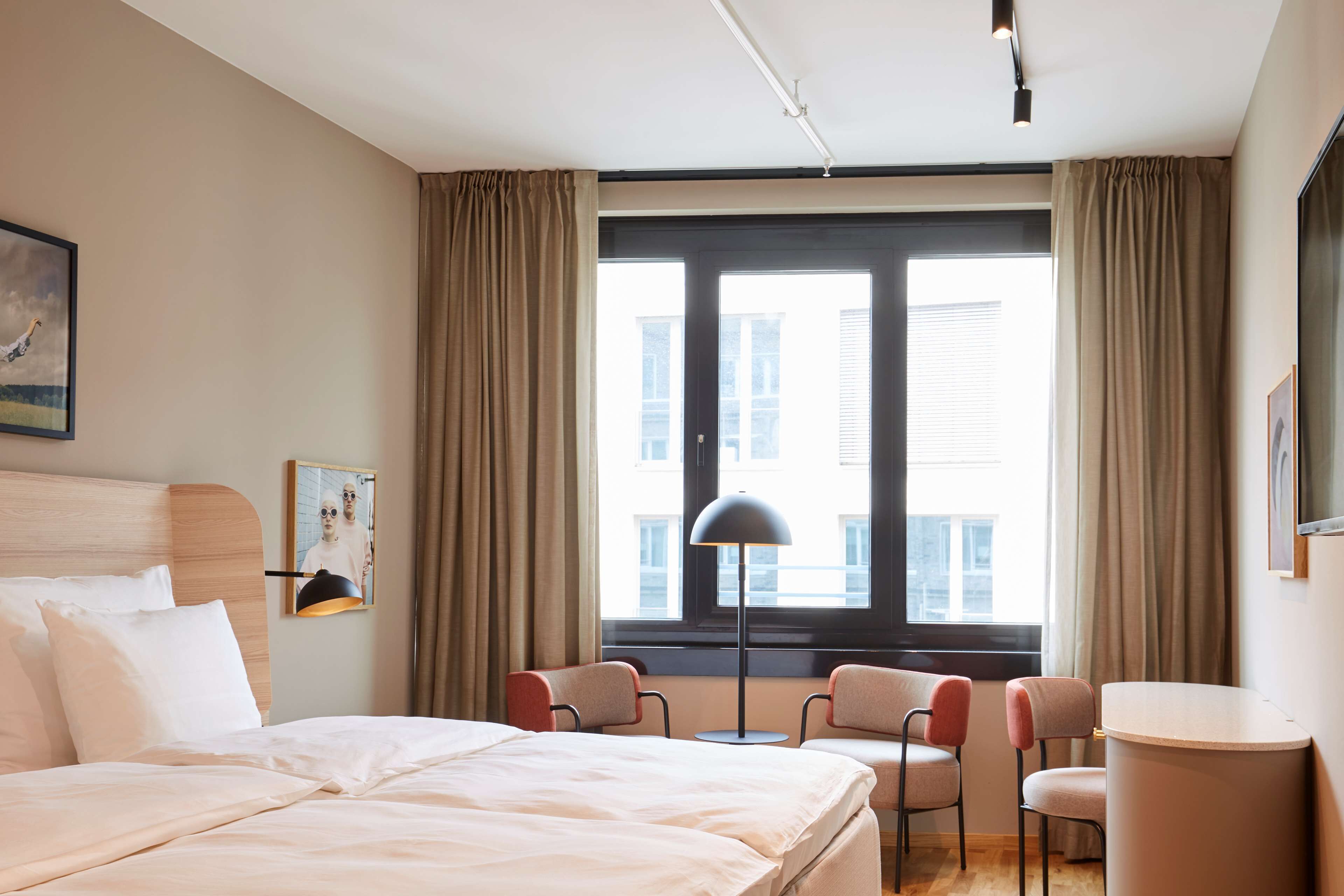 A hotel room with a bed, two chairs, a desk, a lamp, and a large window with curtains.