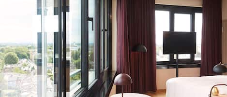 A hotel room with a large window, a bed, a desk, a chair, and a TV.