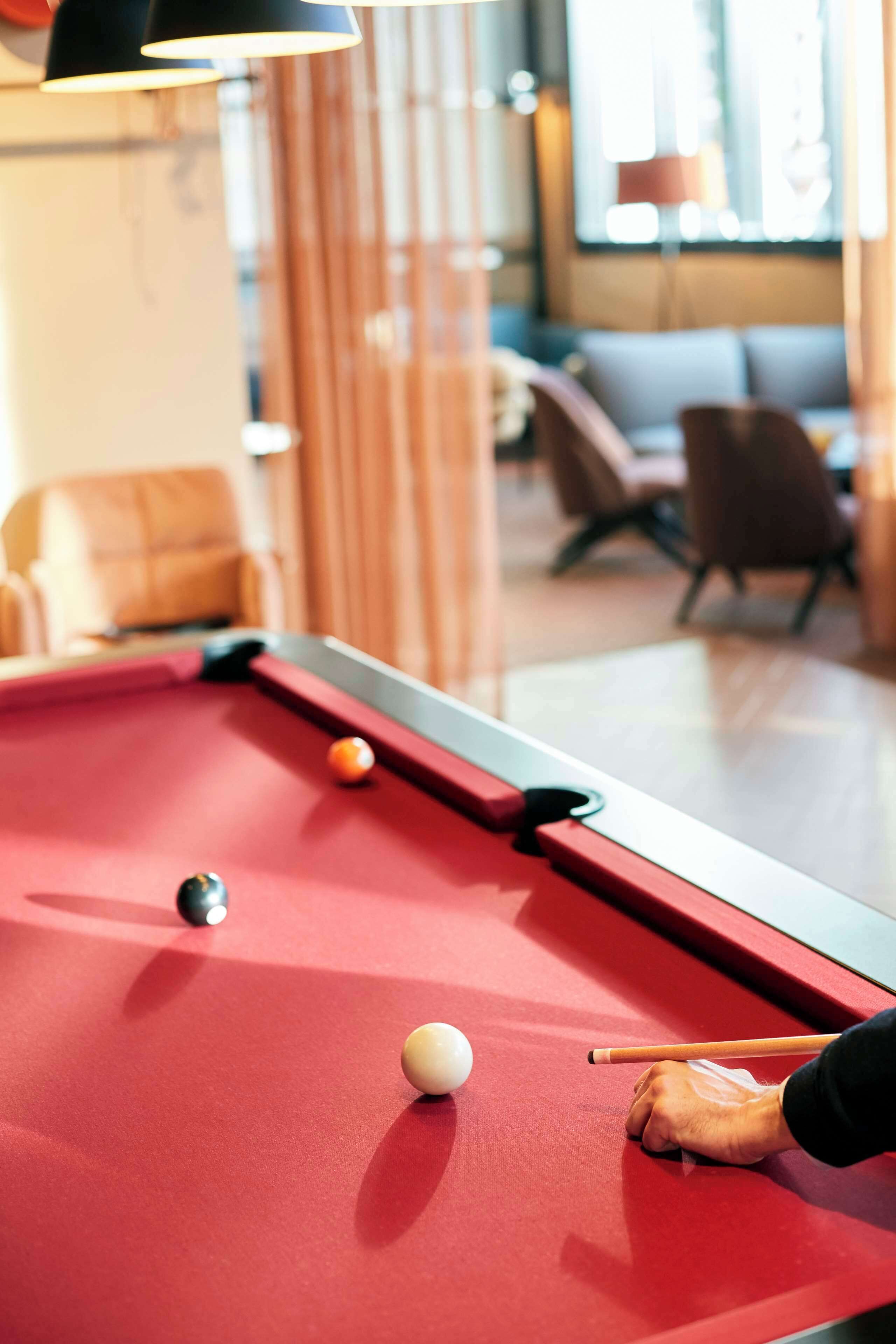 A pool table with balls and a cue stick, surrounded by a modern room with seating and a window.