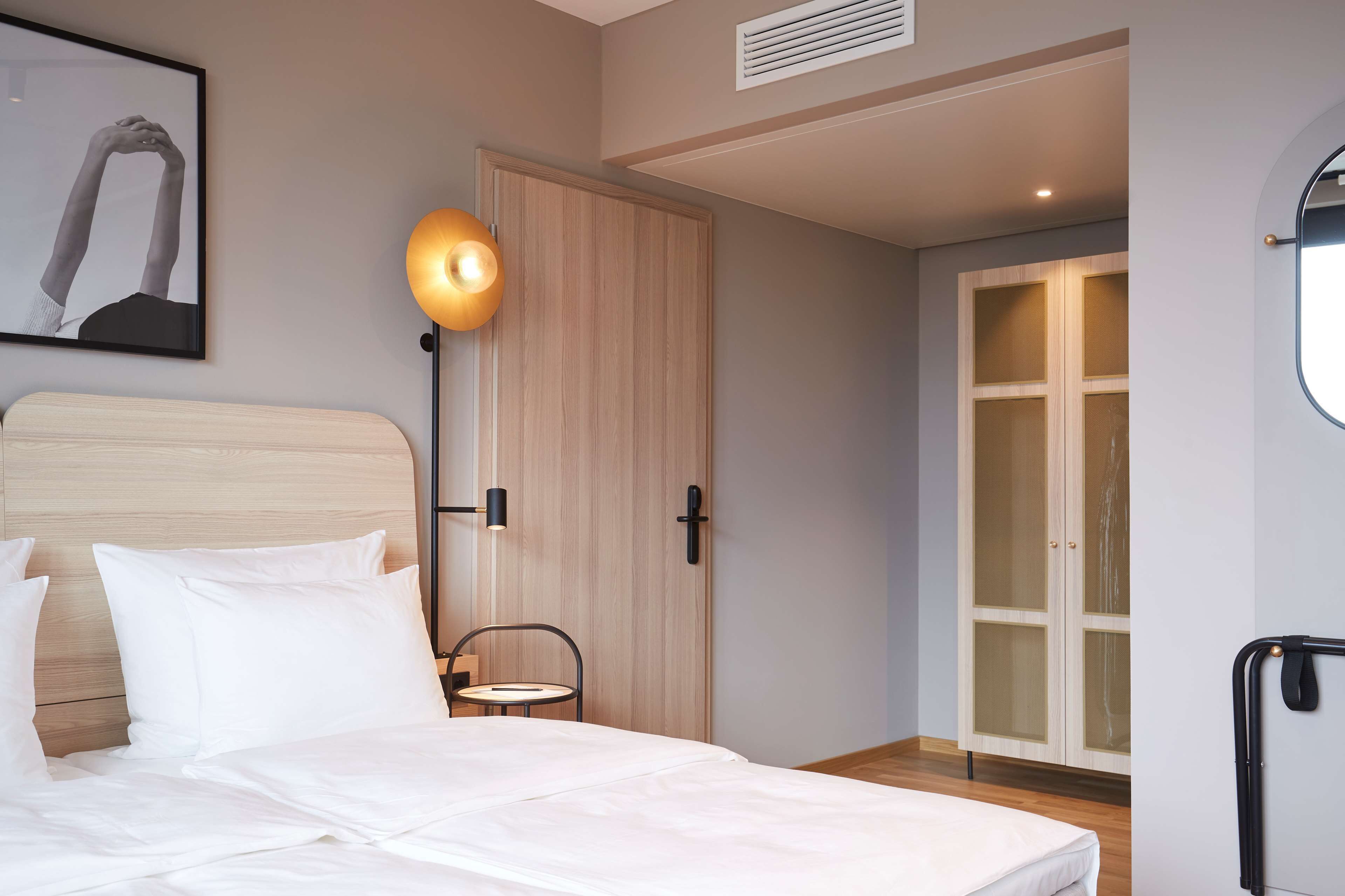 A neatly made bed with white linens, a framed picture on the wall, a bedside lamp, a wooden wardrobe, and a sliding glass door leading to a balcony.