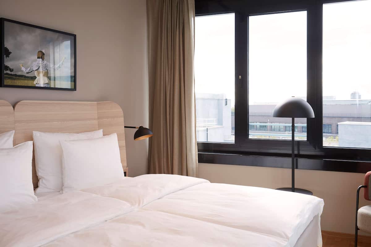 Hotellihuone, jossa on suuri sänky, taulu seinällä, ikkuna, josta on näkymä kaupunkiin, ja lattiavalaisin.